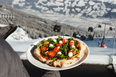 Hand hält Teller mit Pizza belegt mit Rucola, Kirschtomaten und Mozzarellakugeln vor verschneiter Berglandschaft mit Gondelbahn