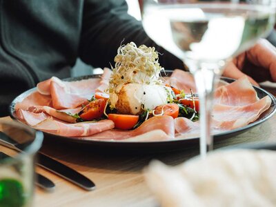 Vorspeisenteller mit Schinken, Tomaten und einem mit Sprossen garnierten Ei, im Vordergrund unscharfes Wasserglas und Besteck