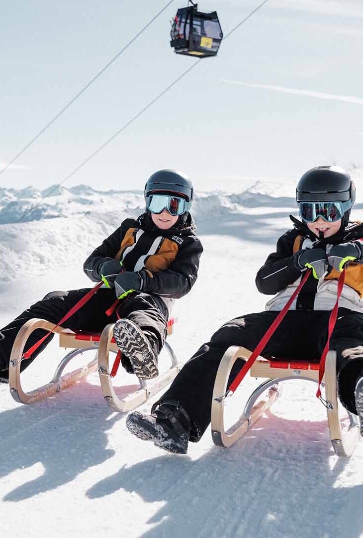 Zwei Personen mit Helm und Winterkleidung sitzen auf Schlitten auf einer verschneiten Piste unter einer Gondelbahn