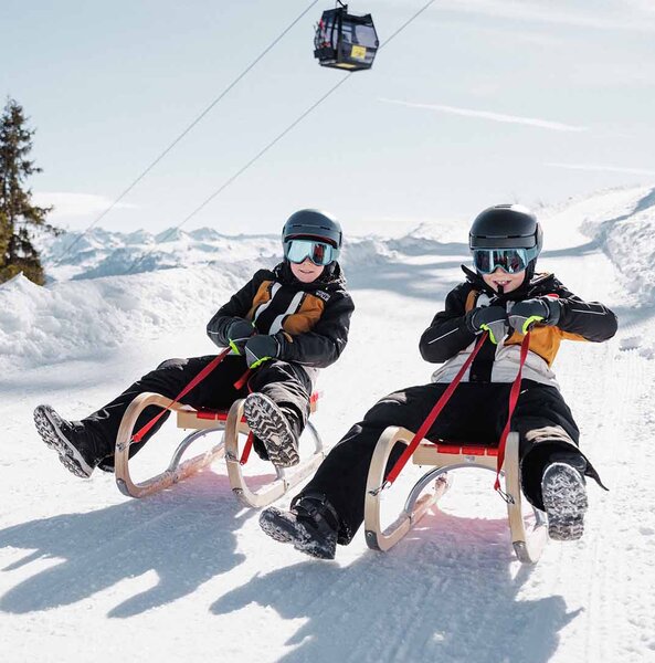 Zwei Personen mit Helm und Winterkleidung sitzen auf Schlitten auf einer verschneiten Piste unter einer Gondelbahn