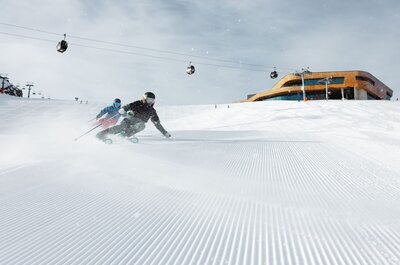 Zwei Skifahrer fahren auf einer frisch präparierten Piste mit Gondeln und einer Bergstation im Hintergrund