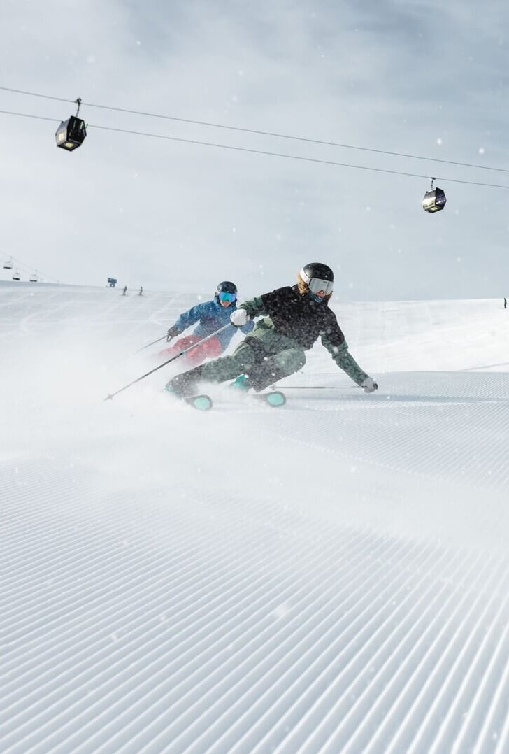 Zwei Skifahrer fahren auf einer frisch präparierten Piste mit Gondeln und einer Bergstation im Hintergrund