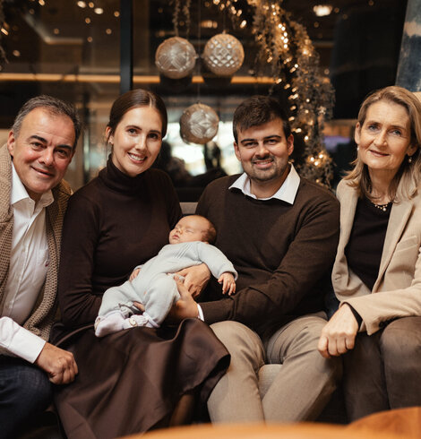 Vier Erwachsene sitzen auf einer Couch, eine Person hält ein schlafendes Baby im Arm, im Hintergrund Weihnachtsdekoration mit Lichterketten und Kugeln
