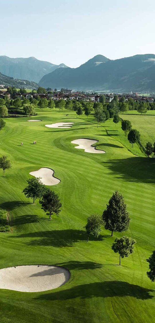 Blick auf eine grüne Golfanlage mit mehreren Sandbunkern, umgeben von Bäumen und Bergen im Hintergrund