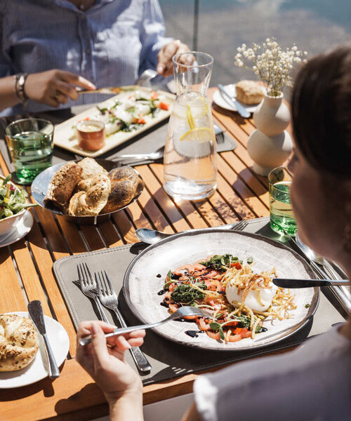 Personen sitzen an einem Holztisch im Freien und essen verschiedene Gerichte aus Tellern und Schalen