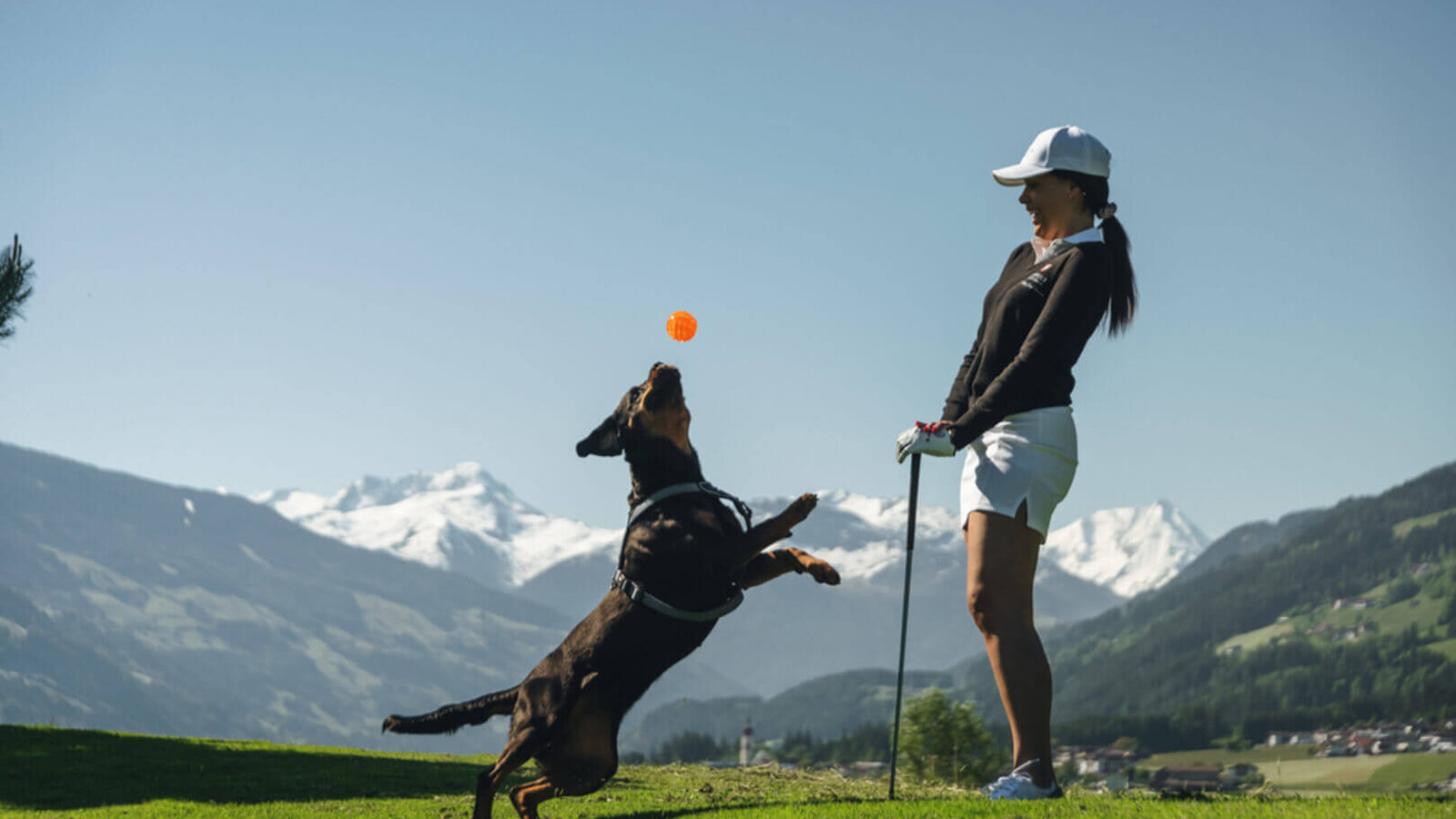 Frau mit Baseballkappe und Shorts wirft Ball für springenden Hund auf einer Wiese vor Bergkulisse