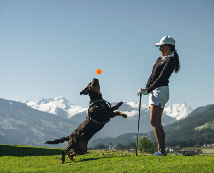 Frau mit Baseballkappe und Shorts wirft Ball für springenden Hund auf einer Wiese vor Bergkulisse