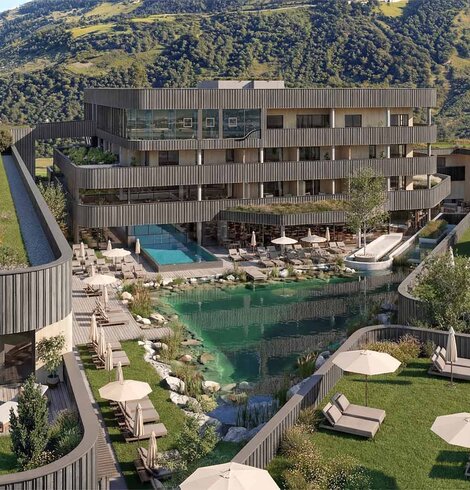 Mehrstöckiges modernes Hotelgebäude mit Holzfassade vor bewaldeten Berg und einem natürlichen Schwimmteich mit Liegestühlen und Sonnenschirmen auf einer Wiese