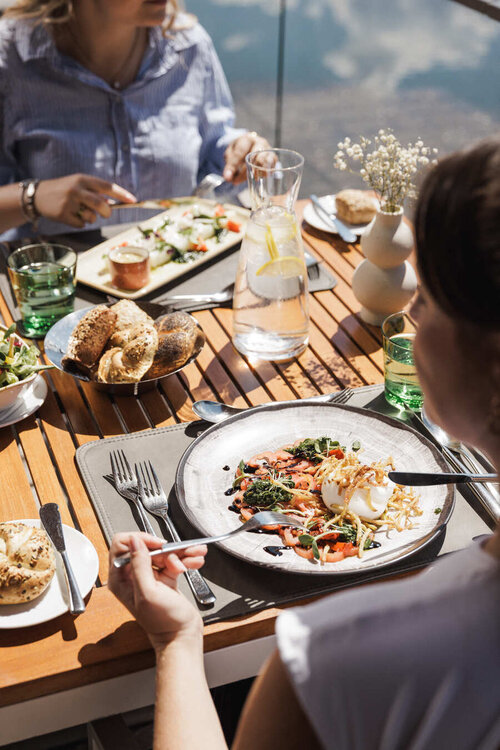 Personen sitzen an einem Holztisch im Freien und essen verschiedene Gerichte aus Tellern und Schalen