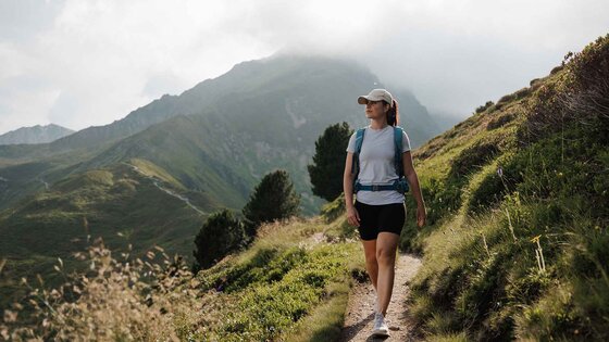 Frau mit Rucksack und Baseballkappe wandert auf schmalem Pfad in bergiger Landschaft mit bewachsenen Hügeln und Bäumen