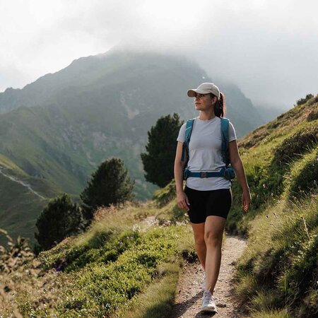Frau mit Rucksack und Baseballkappe wandert auf schmalem Pfad in bergiger Landschaft mit bewachsenen Hügeln und Bäumen