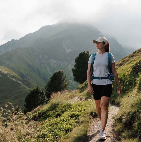 Frau mit Rucksack und Baseballkappe wandert auf schmalem Pfad in bergiger Landschaft mit bewachsenen Hügeln und Bäumen