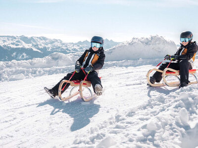Zwei Kinder mit Helmen und Skibrillen sitzen auf Holzschlitten auf einer verschneiten Piste vor Bergpanorama