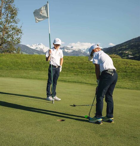 Zwei Kinder auf einem Golfplatz eines hält die Fahne das andere schlägt den Ball