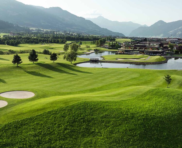 Landschaft mit Golfplatz, Teich, Bäumen und Bergen im Hintergrund