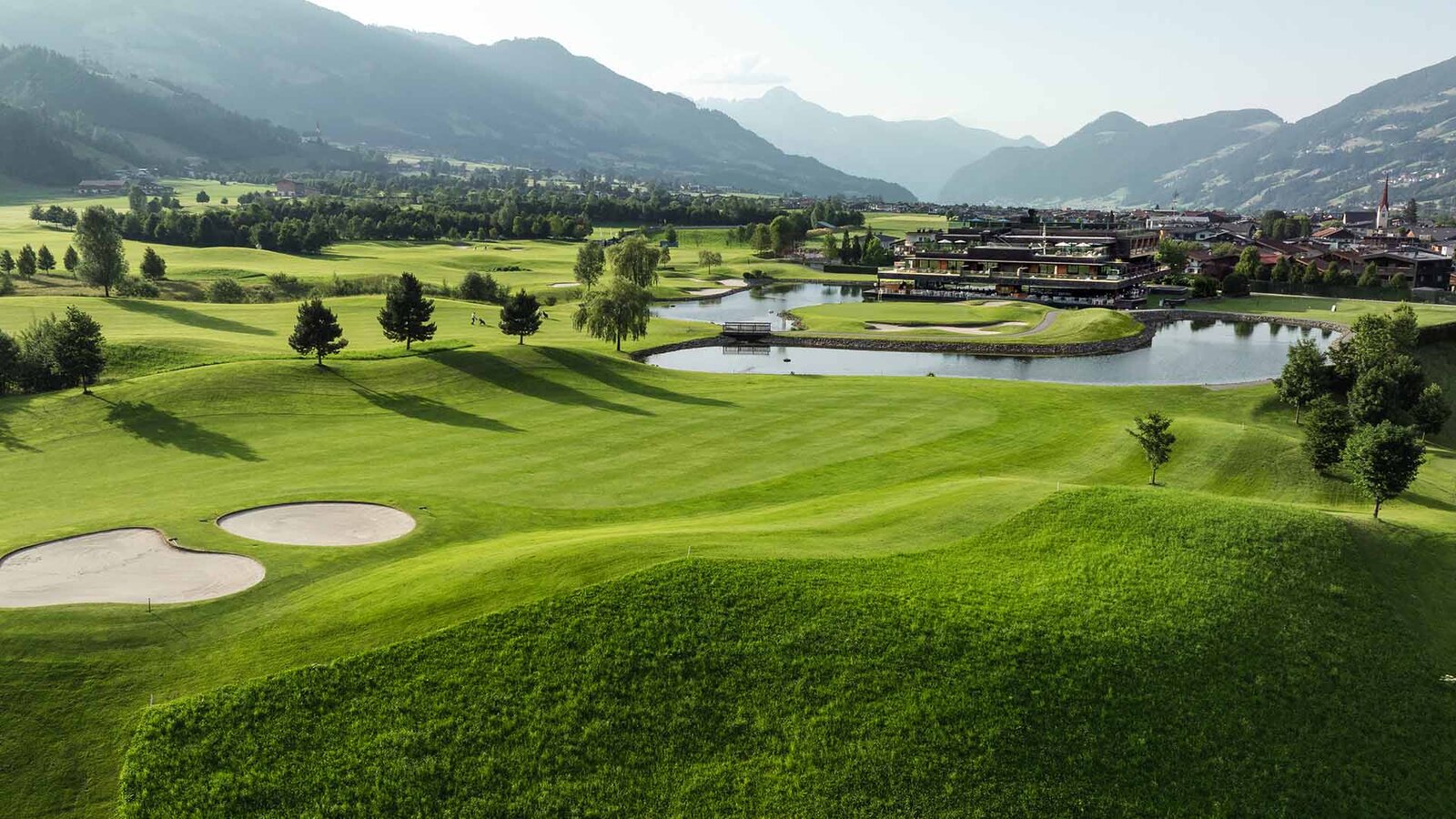 Landschaft mit Golfplatz, Teich, Bäumen und Bergen im Hintergrund
