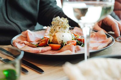 Vorspeisenteller mit Schinken, Tomaten und einem mit Sprossen garnierten Ei, im Vordergrund unscharfes Wasserglas und Besteck