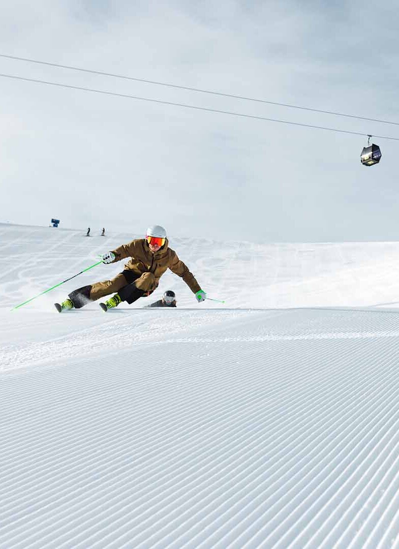 Skifahrer in leichter Hocke fährt auf frisch präparierter Piste mit Seilbahn und modernem Gebäude im Hintergrund