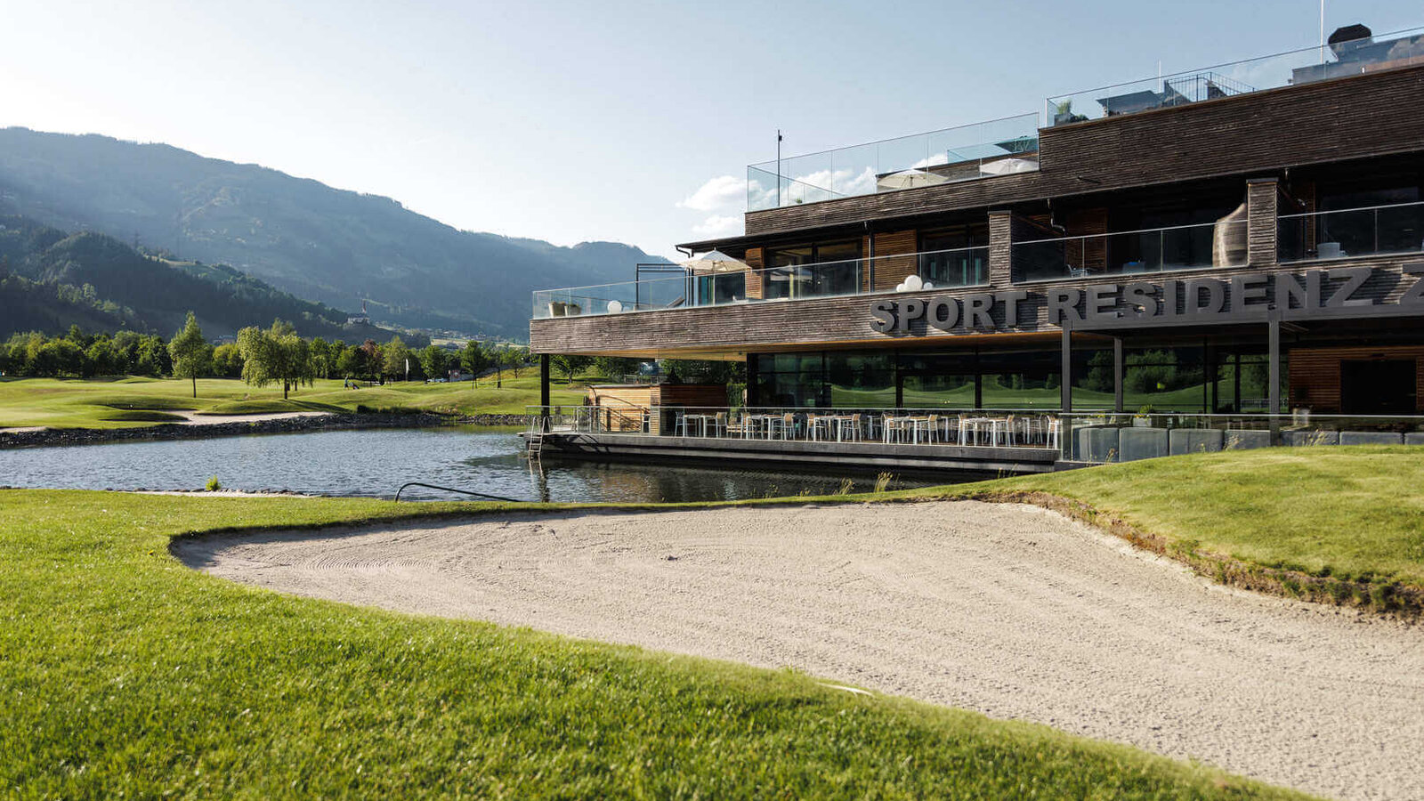 Sportresidenz in seitlicher Ansicht mit See im Vordergrund und Bergen im Hintergrund