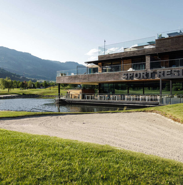 Sportresidenz in seitlicher Ansicht mit See im Vordergrund und Bergen im Hintergrund