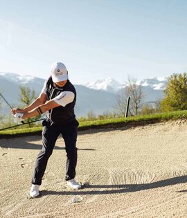 Golfer in Schwungposition im Sandbunker mit Golftasche im Hintergrund und Berglandschaft