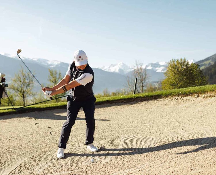 Golfer in Schwungposition im Sandbunker mit Golftasche im Hintergrund und Berglandschaft