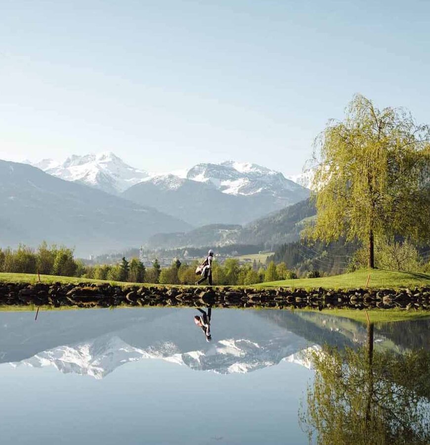 Spiegelglatter See mit Bäumen am Ufer und schneebedeckten Bergen im Hintergrund