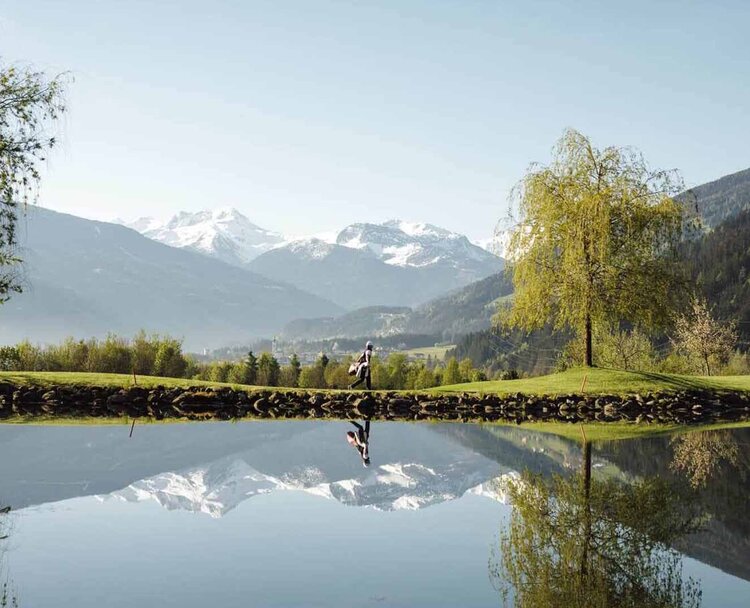Spiegelglatter See mit Bäumen am Ufer und schneebedeckten Bergen im Hintergrund