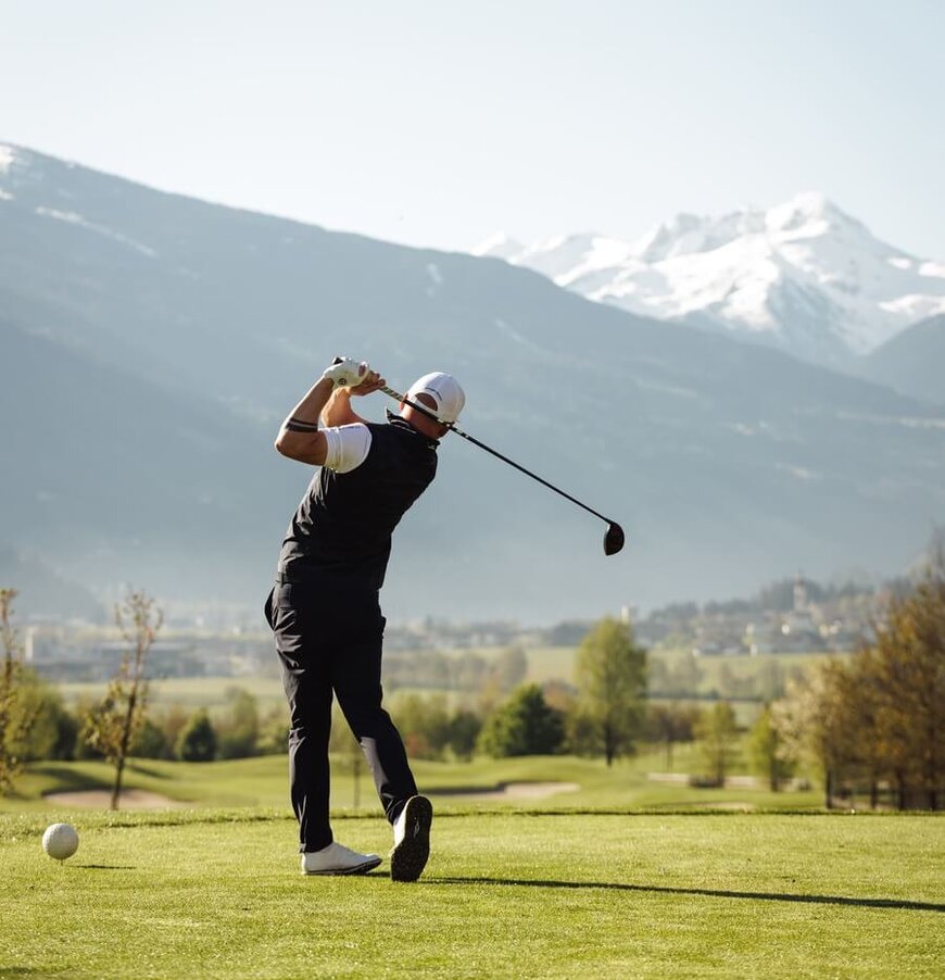 Golfer in schwarzer Kleidung beim Abschlag auf einem Golfplatz mit Bergen im Hintergrund