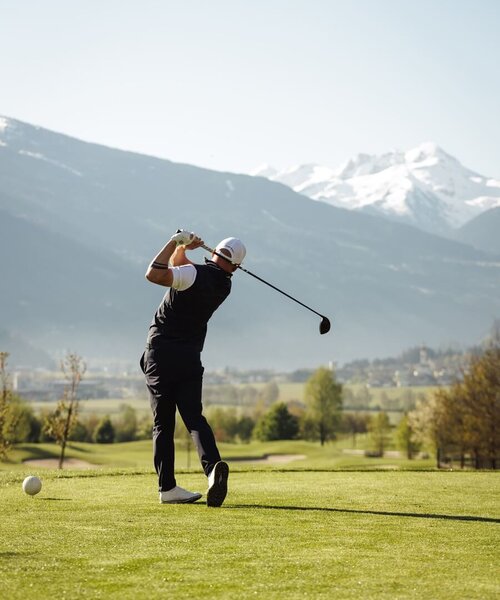 Golfer in schwarzer Kleidung beim Abschlag auf einem Golfplatz mit Bergen im Hintergrund