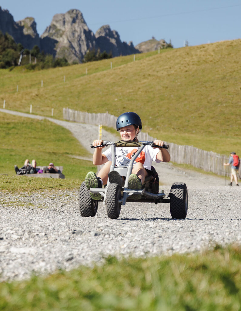 Person mit Helm sitzt auf einem vierrädrigen Tretfahrzeug auf einem Schotterweg in bergiger Landschaft