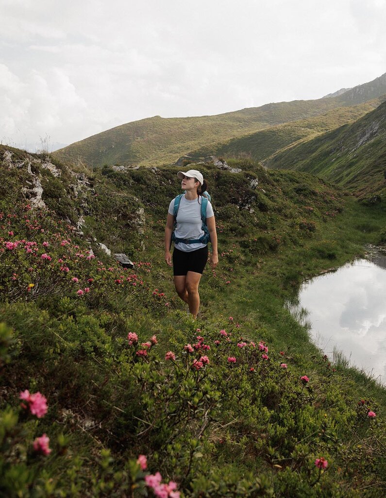 Wanderin mit Rucksack und Kappe geht auf einem Pfad entlang eines Bergsees in einer grünen Berglandschaft