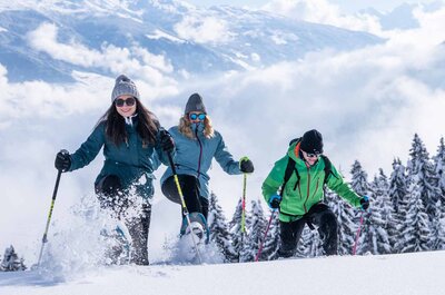 Drei Personen mit Winterkleidung und Skistöcken laufen durch tiefen Schnee vor verschneiten Tannenbäumen unter blauem Himmel mit Wolken