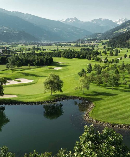 Blick auf eine grüne Golfanlage mit Teich, Bunkern und Bergen im Hintergrund