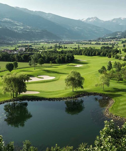 Blick auf eine grüne Golfanlage mit Teich, Bunkern und Bergen im Hintergrund