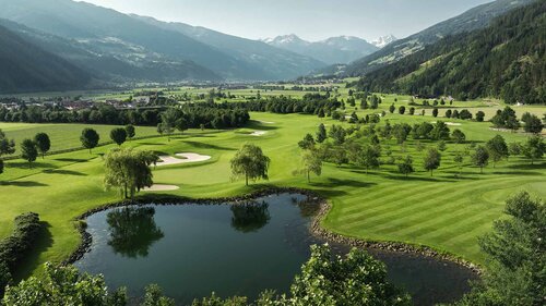 Blick auf eine grüne Golfanlage mit Teich, Bunkern und Bergen im Hintergrund