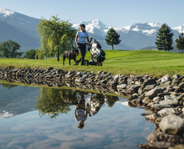 Person mit Golfwagen auf einem Golfplatz vor schneebedeckten Bergen und Spiegelung im Wasser