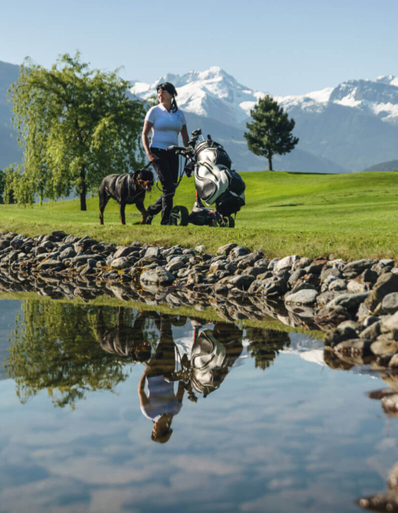 Person mit Golfwagen auf einem Golfplatz vor schneebedeckten Bergen und Spiegelung im Wasser