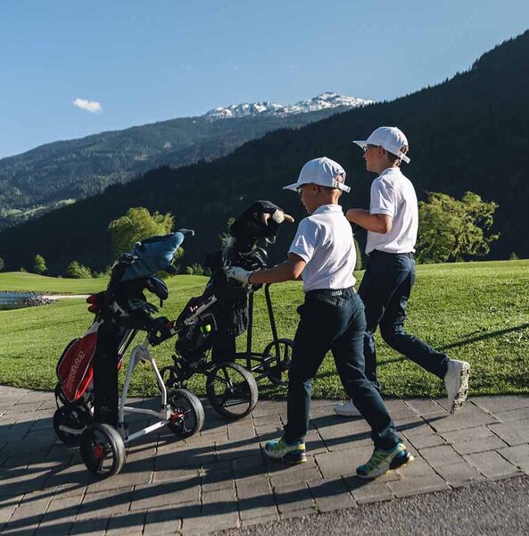 Zwei Jungen in Golfkleidung schieben Golfwagen auf einem Weg neben einem Golfplatz mit Bergen im Hintergrund