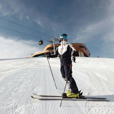 Skifahrer mit Helm und Skibrille steht auf präparierter Skipiste vor einer Bergstation und einer Gondelbahn unter bewölktem Himmel