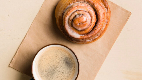 Kaffeetasse mit Schaum neben einer Zimtschnecke auf hellem Untergrund