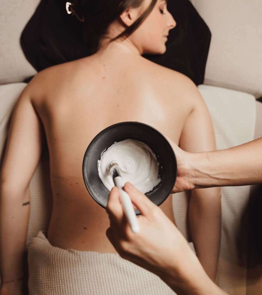 Rücken einer liegenden Person mit einem schwarzen Schälchen und einem Pinsel, der weiße Creme aufträgt