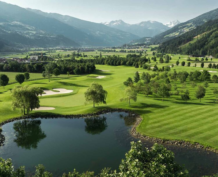 Landschaft mit Golfplatz, Teich, Bäumen und Bergen im Hintergrund